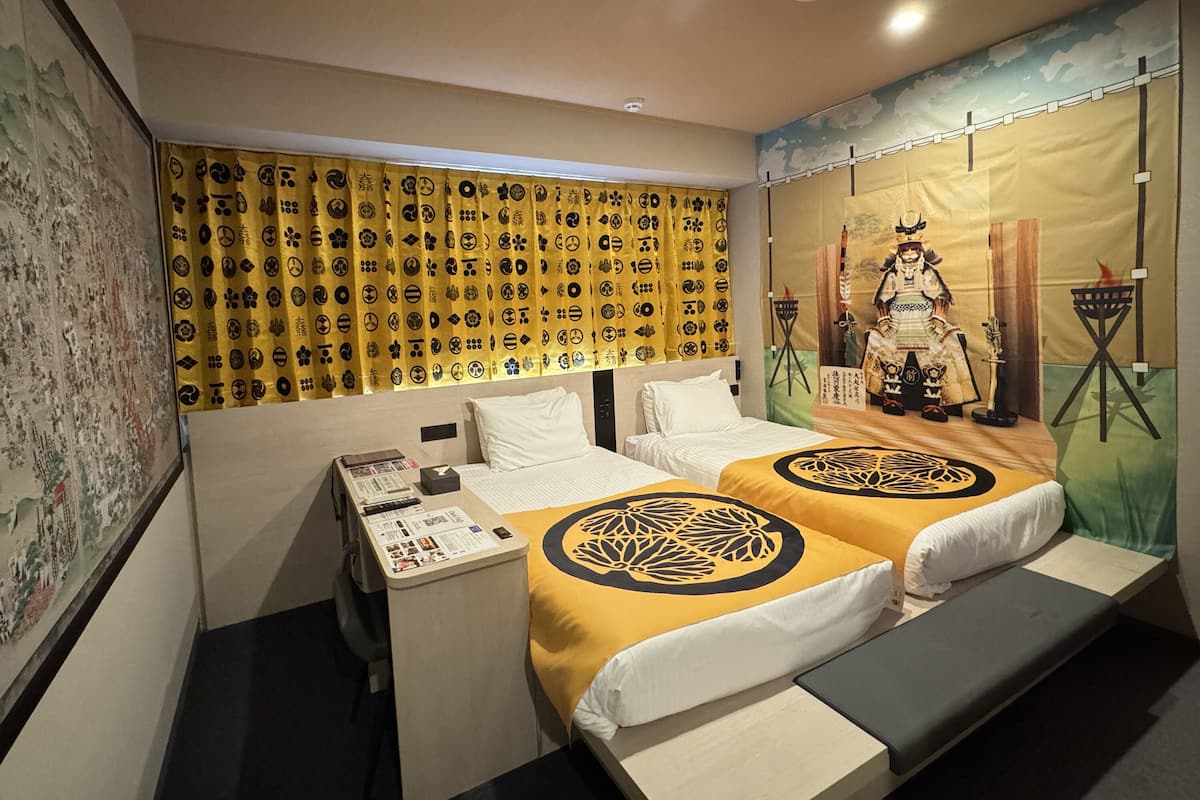 A hotel room with two beds, a desk with a phone and a small table, and a mural on the wall.