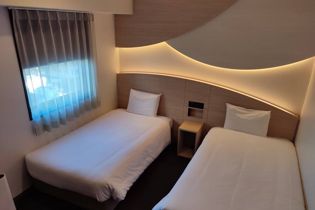 A modern hotel room with two single beds, a bedside table, and a window with sheer curtains.