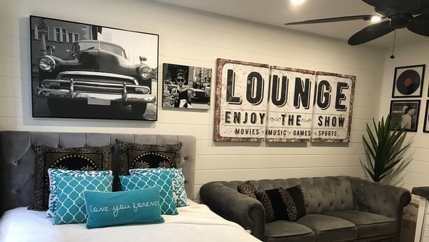 Une chambre avec un lit, un canapé gris et un panneau mural sur lequel on peut lire : « RELAXEZ-VOUS, PROFITEZ DES FILMS, DE LA MUSIQUE, DES JEUX ET DES ÉMISSIONS SPORTIVES ».