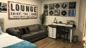 Une pièce chaleureuse avec un canapé gris, un bureau avec des tiroirs, un ventilateur de plafond et un mur orné de pochettes de disques encadrées, ainsi qu'une pancarte sur laquelle on peut lire : « ESPACE DÉTENTE – PROFITEZ DES FILMS, DE LA MUSIQUE, DES JEUX ET DES ÉMISSIONS SPORTIVES ».