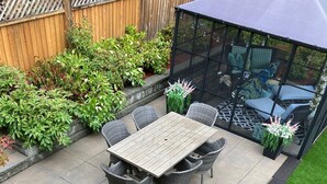Une terrasse avec une table et des chaises en bois, entourée de différentes plantes et protégée par une structure en verre.