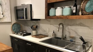 Une cuisine moderne avec des meubles de couleur sombre, un plan de travail blanc, un évier en acier inoxydable, un four à micro-ondes et une bouteille de vin posée sur une étagère.