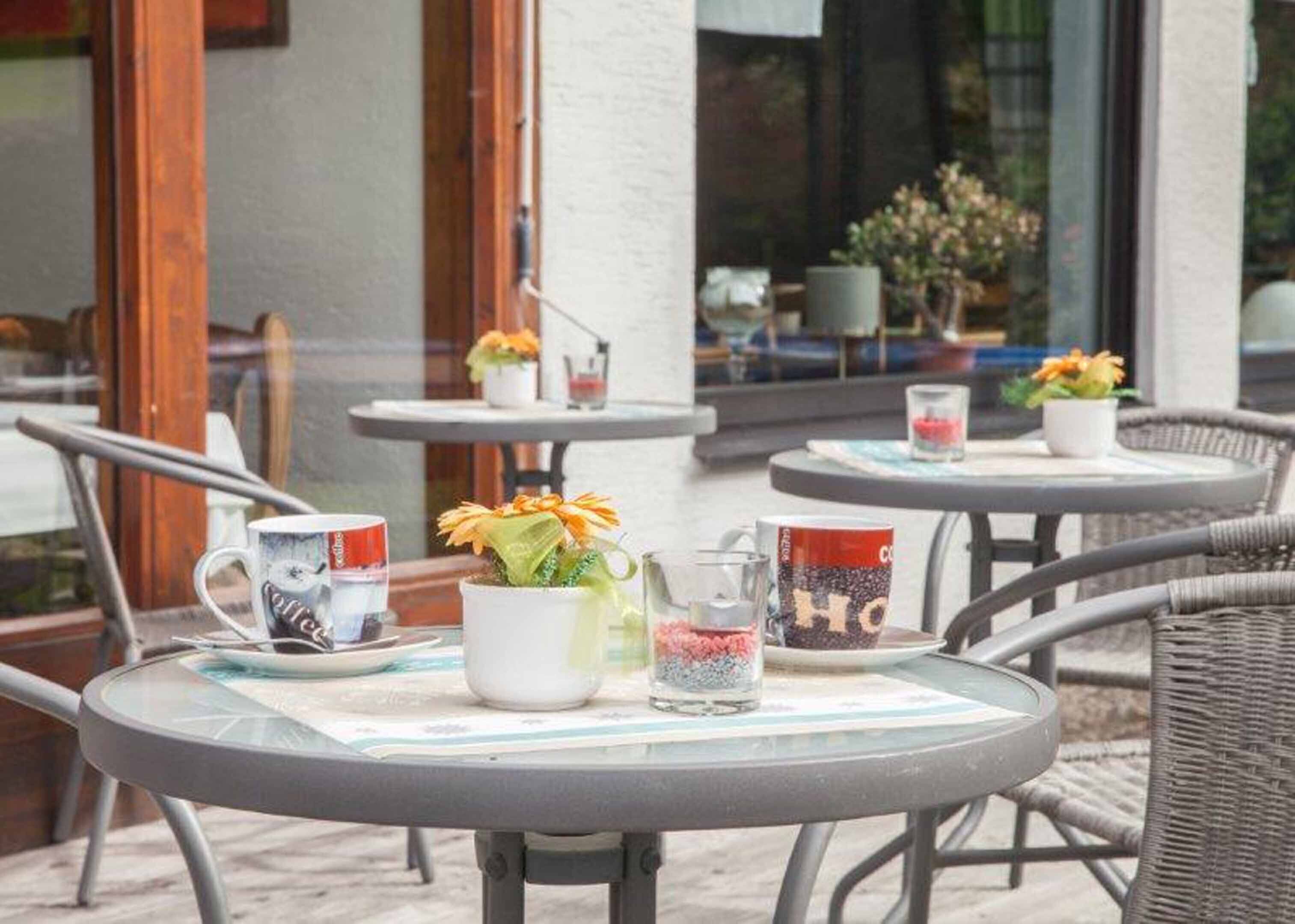 An outdoor seating area with round tables, some with potted plants and coffee mugs.
