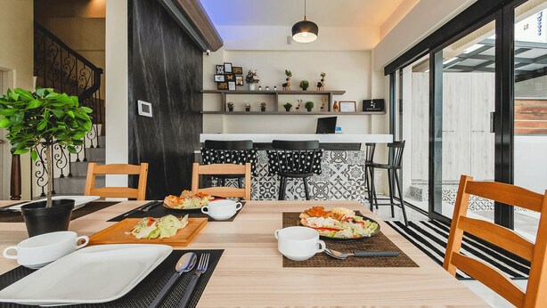 Un coin repas avec une table en bois dressée pour deux personnes, un mur orné d’un motif noir et blanc, et une cuisine en arrière-plan.