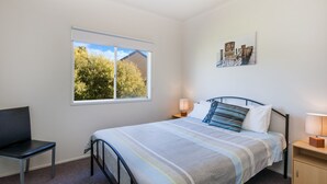 A bedroom with a bed, a chair, a nightstand, a lamp, a window, and a painting on the wall. - Julieanne (Port Fairy)