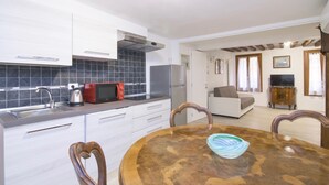 A compact kitchen with white cabinets, a black tiled backsplash, and a red microwave. There's a dining area with a wooden table and chairs, and a living room with a sofa and a TV. - Ca' Alberto Apartments 1 (Venice)
