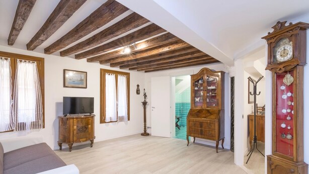 A spacious living room with exposed wooden beams, a large wooden cabinet with glass doors, a television, and a large wall clock. - Ca' Alberto Apartments 1 (Venice)