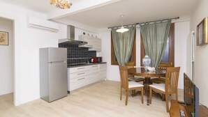 A compact kitchen with a refrigerator, stove, and modern cabinetry. There is a dining area with a wooden table and chairs, and a window with curtains. - Ca' Alberto Apartments 2 (Venice)
