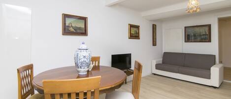 A dining area with a round wooden table, chairs, a vase, and a television. There is a grey sofa and a painting on the wall.