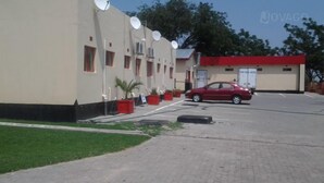 Un edificio con antenas parabólicas, un coche rojo estacionado adelante y un pequeño jardín con macetas.