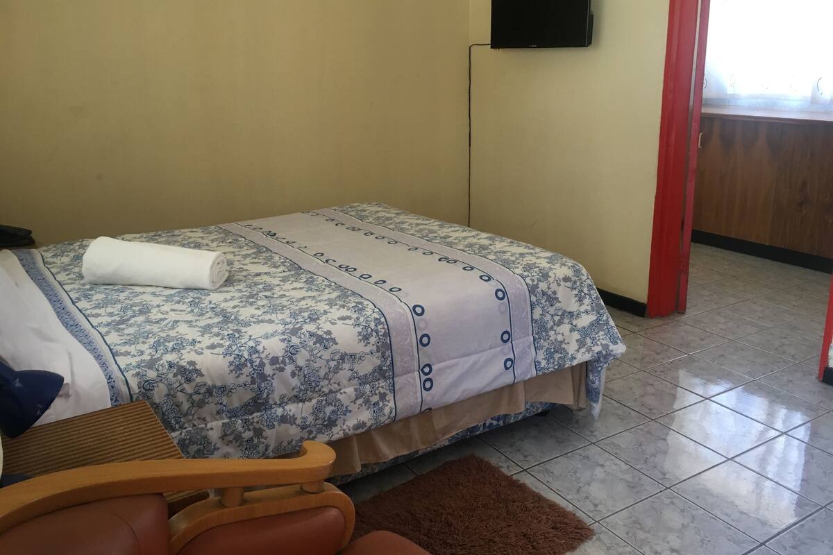 Ein Hotelzimmer mit einem Bett, einem Holzstuhl, einem braunen Teppich und einem an der Wand befestigten Fernseher.