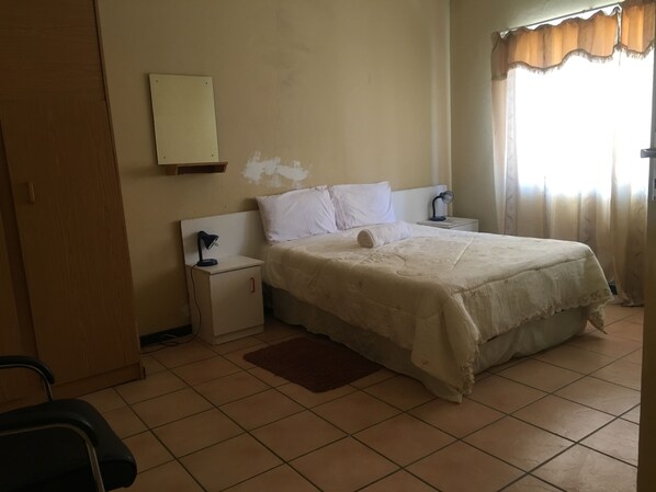 A bedroom with a bed, nightstands, a desk lamp, a mirror, and a window with curtains. - Tshesebe Stopover Motel (Francistown)