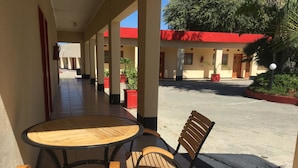 Un área de estar al aire libre techada, con una mesa redonda y dos sillas de madera, piso de baldosas y un edificio con tejado rojo al fondo.