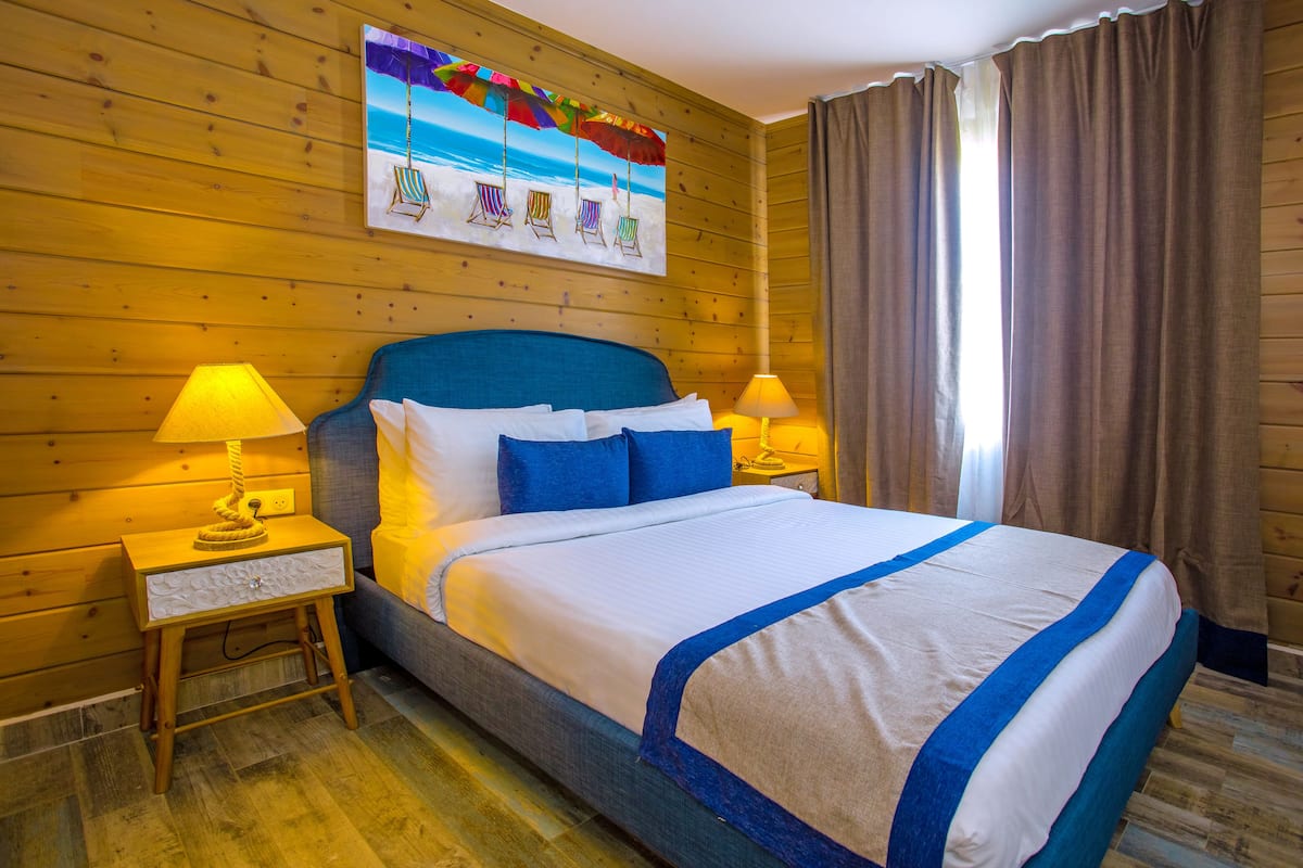 Une chambre d’hôtel avec un décor intérieur en bois, un grand lit avec une literie bleue, deux lampes de chevet et un tableau sur le thème de la plage accroché au mur.