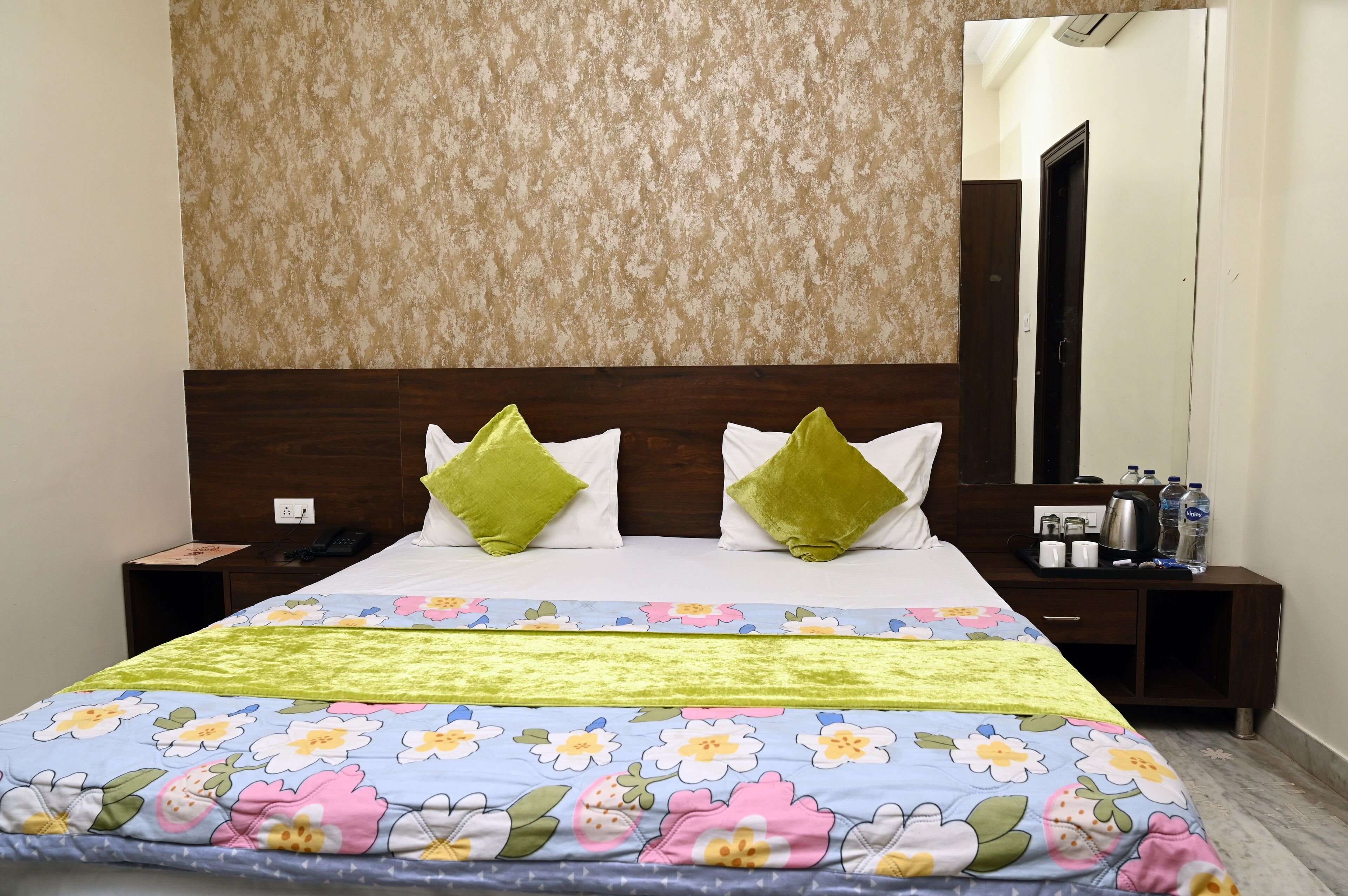Une chambre d’hôtel avec une couvre-lit à motifs floraux, une tête de lit en bois, deux coussins verts, une table de chevet avec un téléphone et un miroir au mur.