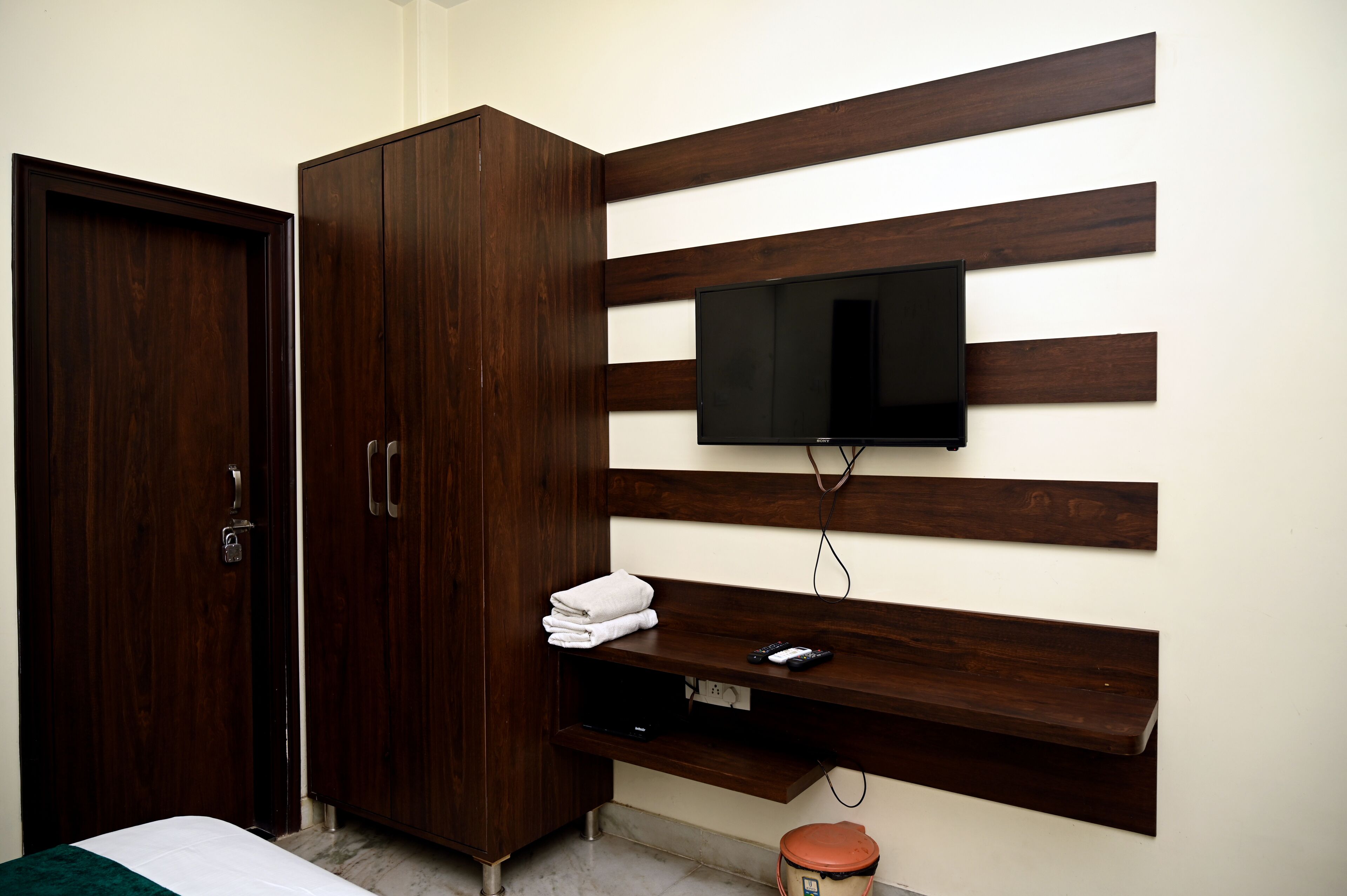 Un dormitorio con un armario de madera, un televisor de pantalla plana montado en una pared de paneles de madera, una mesita de noche con un control remoto y un pequeño cubo de basura.