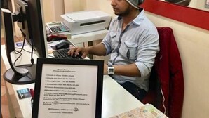 Ein Mann sitzt am Schreibtisch mit einem Computer, einem Drucker und einem gerahmten Dokument.