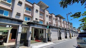 Una hilera de edificios de varios pisos con tiendas en la planta baja, algunas con persianas cerradas, y un cielo azul despejado arriba.