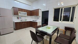 Una cocina moderna con azulejos blancos, armarios de madera, un refrigerador de acero inoxidable, un microondas integrado y un comedor con una mesa y sillas de vidrio.