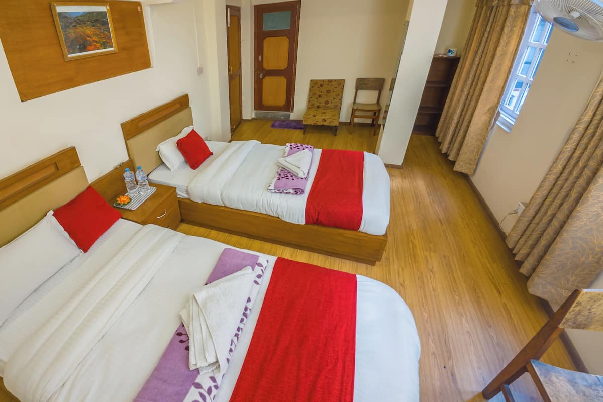 A hotel room with two beds, wooden headboards, a small table with water bottles, a chair, and a window with curtains.