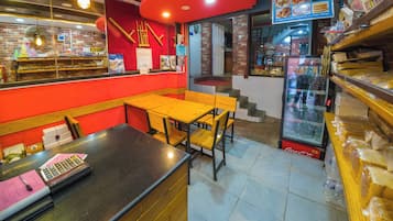 A restaurant interior with wooden tables and chairs, a cash register, and a refrigerator stocked with beverages.