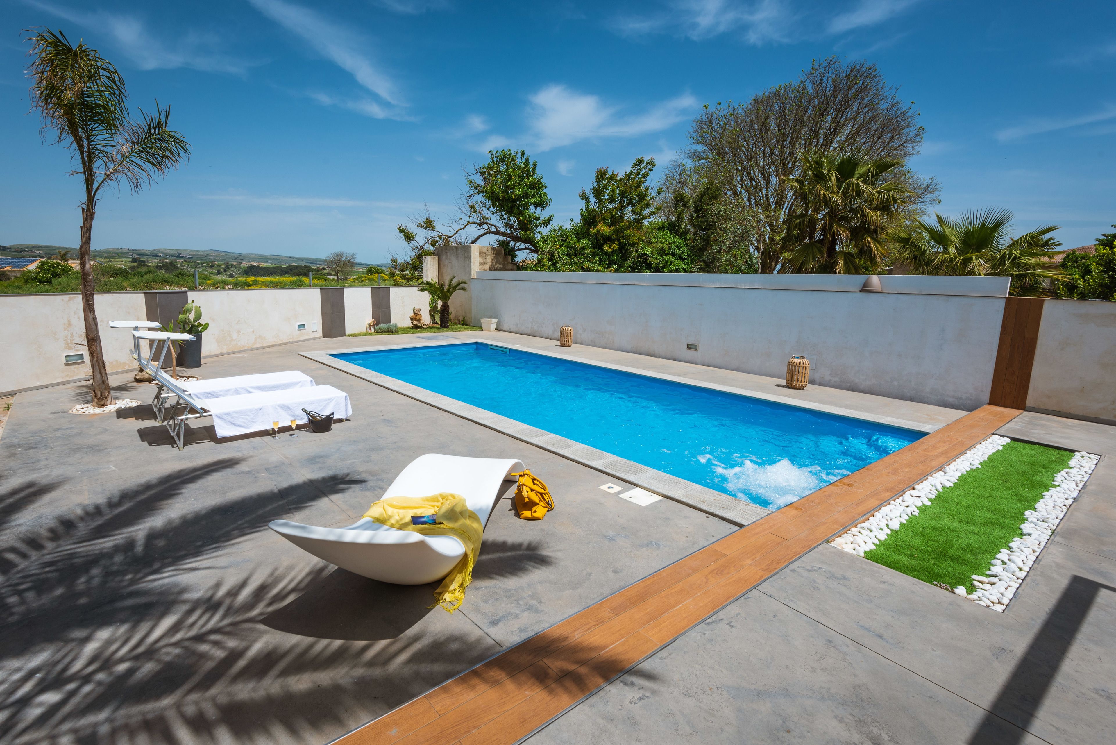 Une piscine entourée d’un petit muret et de verdure, avec des chaises longues et une petite table à proximité.