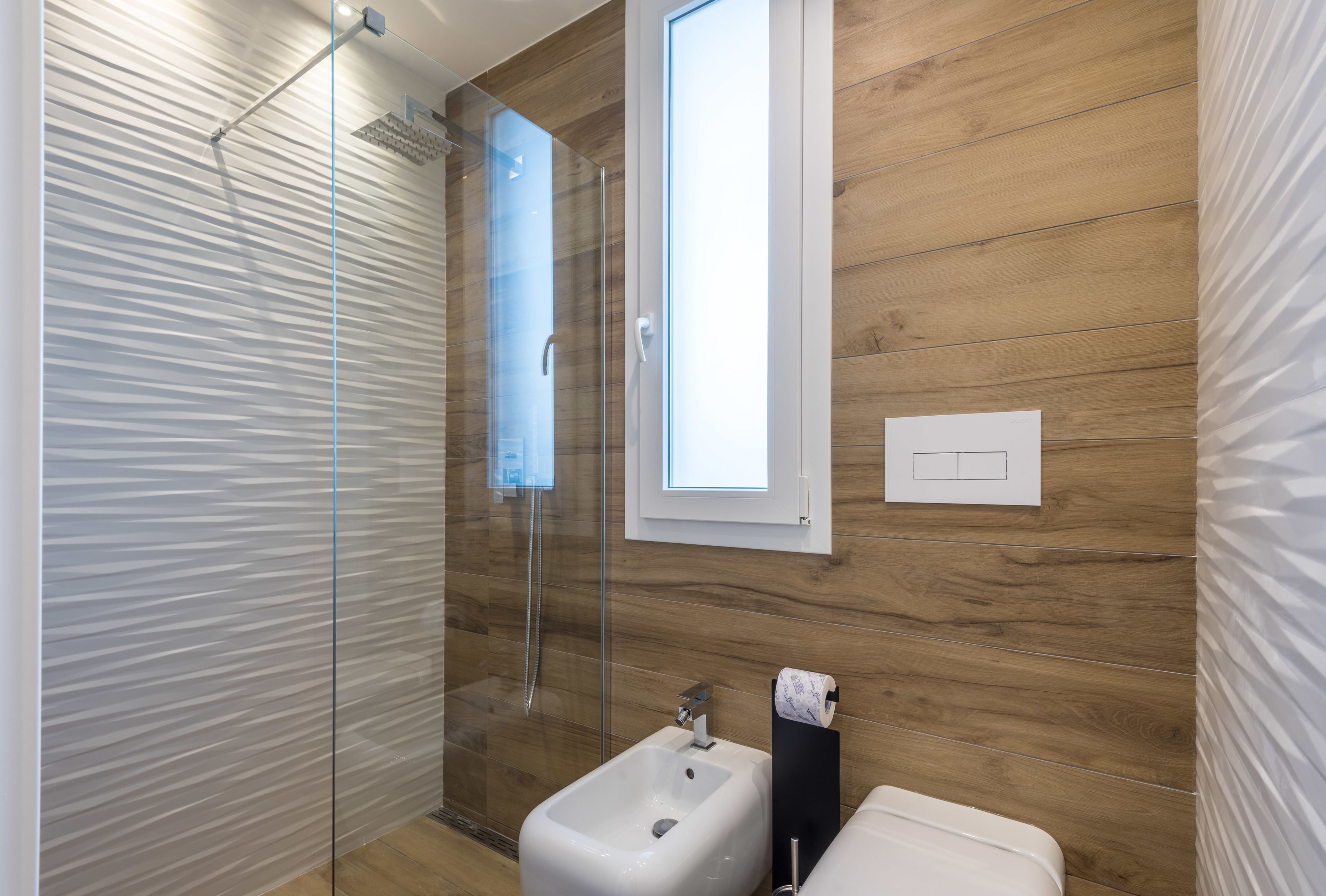 A modern bathroom with a glass shower enclosure, a bidet, and a window providing natural light.