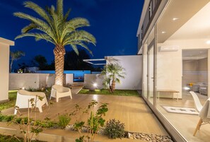 A modern outdoor patio with a palm tree, seating area, and a glass door leading to a room. - Aurienzia (Ragusa)