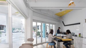 A modern cafe with large windows, a counter with a barista, and a seating area with stools and tables. - TG Hostel (Taichung)