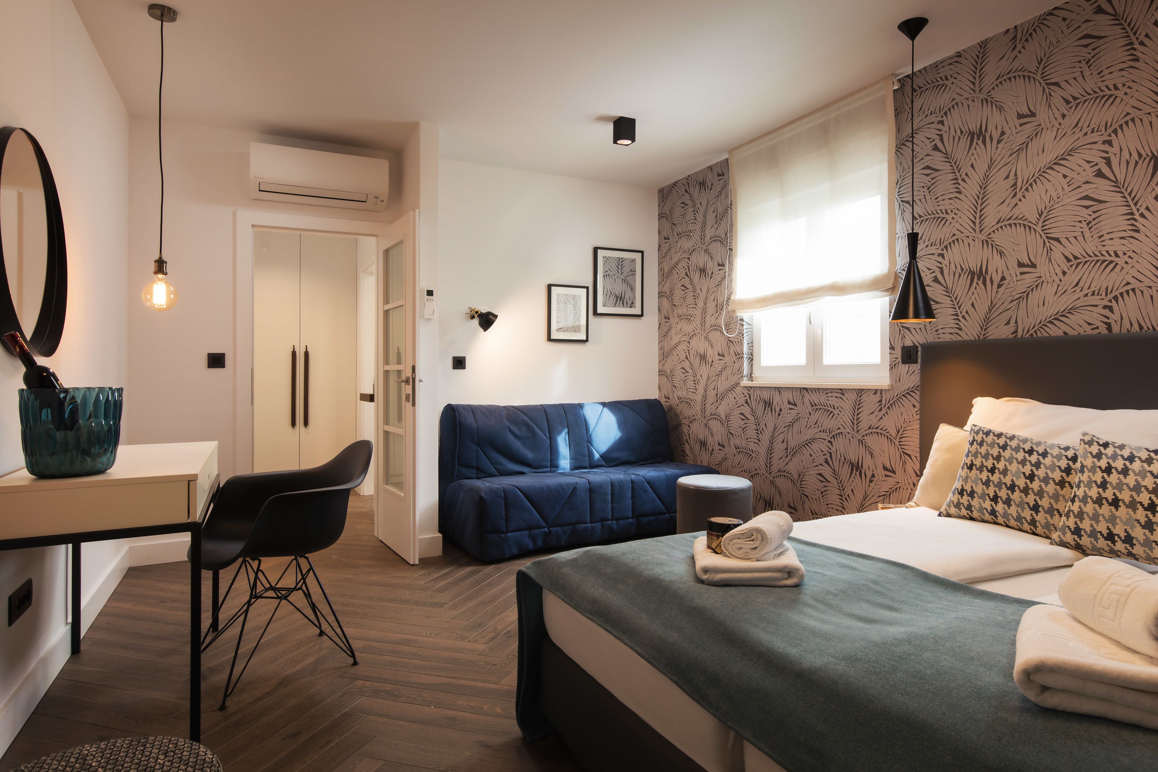 Une chambre d’hôtel moderne équipée d’un lit, d’un bureau, d’une chaise, d’un canapé et d’une fenêtre donnant sur un mur orné de motifs végétaux.