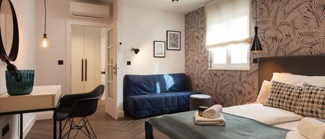 Une chambre d’hôtel moderne équipée d’un lit, d’un bureau, d’une chaise, d’un canapé et d’une fenêtre donnant sur un mur orné de motifs végétaux.
