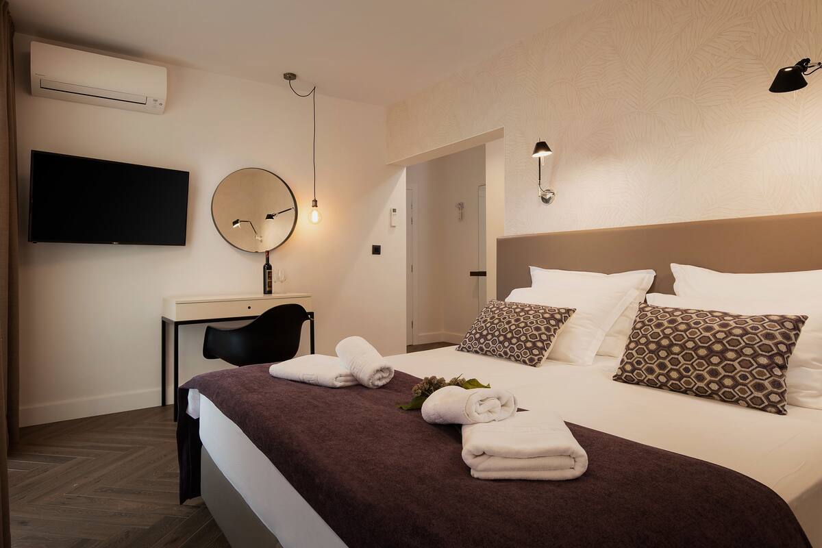 A hotel room with a bed, a desk, a flat-screen TV, a mirror, and a wall-mounted light fixture.