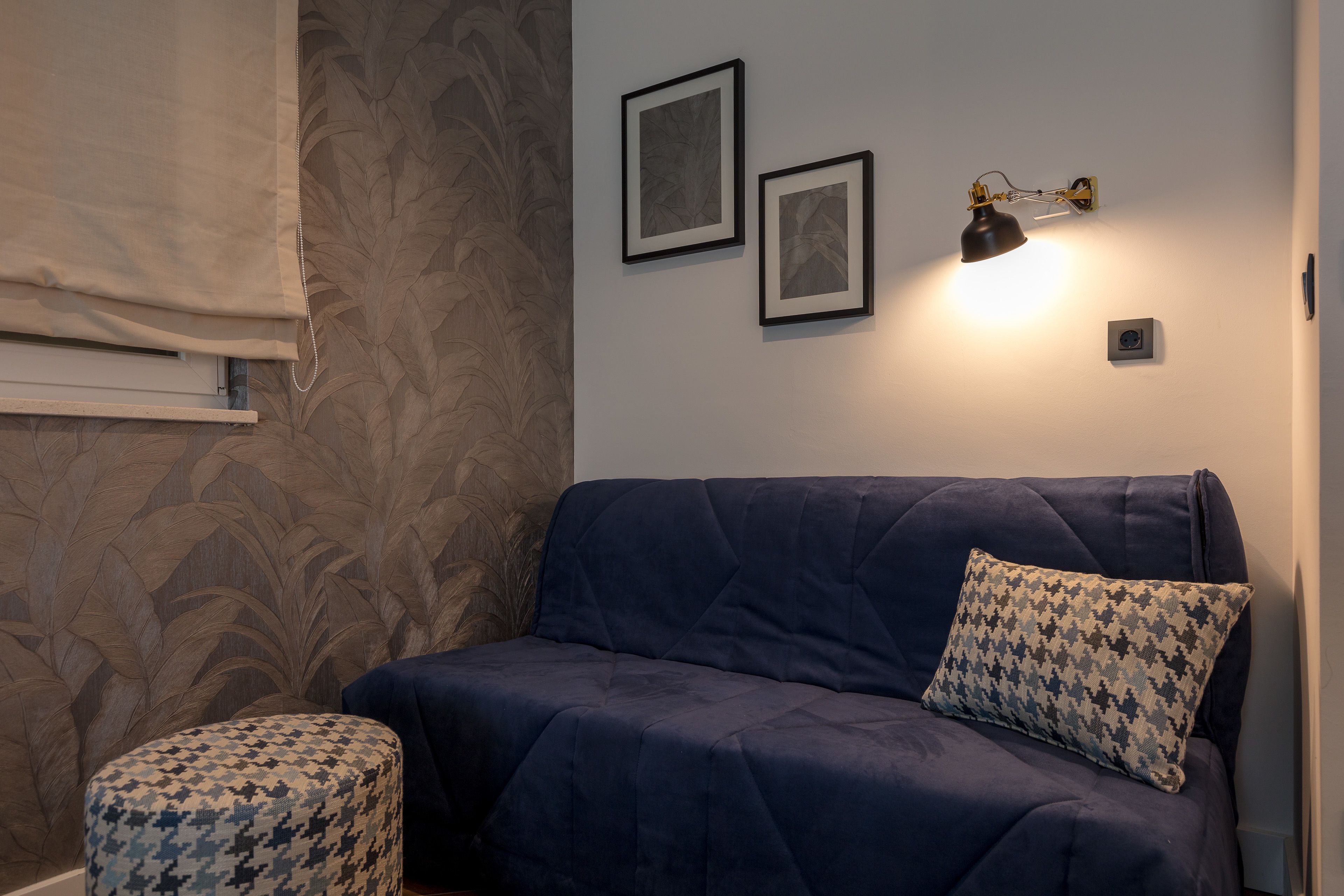 A dark blue sofa with a patterned pillow, a wall-mounted picture frame, and a wall-mounted lamp.