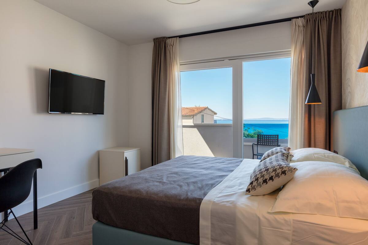 A hotel room with a bed, a TV, a desk, and a view of the sea.