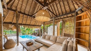 A poolside lounge with a thatched roof, a sofa, a wooden table, and a hanging light fixture. - La Cabane Bali (Jimbaran)