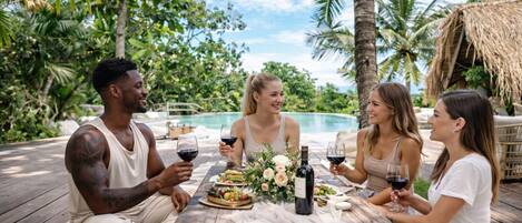 A group of people dining outdoors on a wooden deck with a pool in the background.