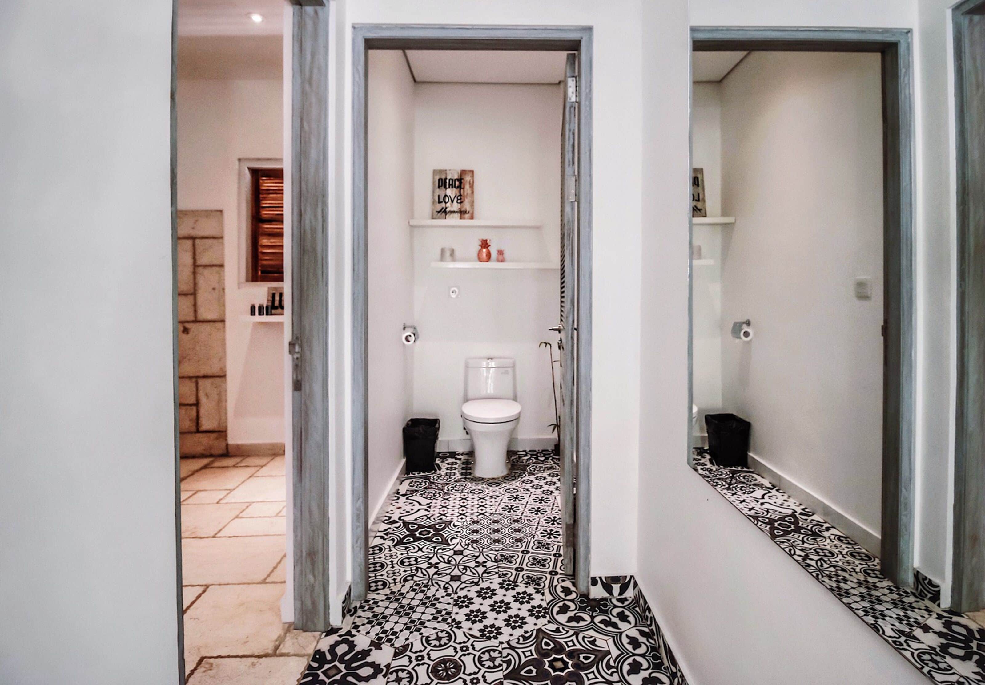 A narrow hallway leading to a bathroom with patterned floor tiles, a toilet, and a shelf with toiletries.