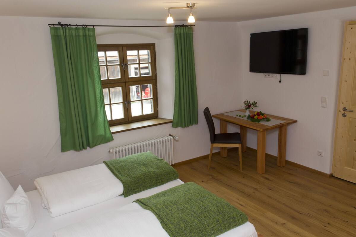 A bedroom with a bed, a desk, a chair, a window with curtains, and a television on the wall.
