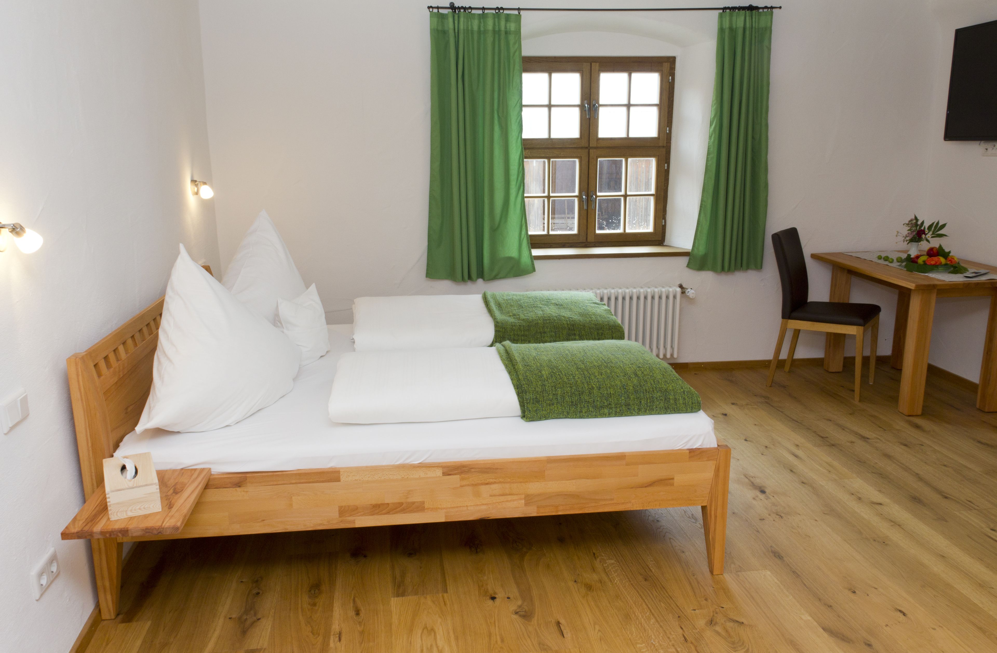 Un lit en bois avec du linge de lit blanc et une couverture verte, un bureau en bois avec une chaise, et une fenêtre avec des rideaux verts.