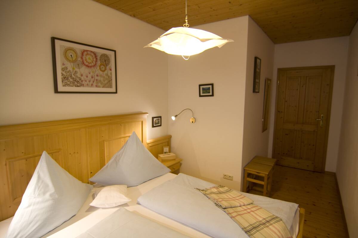 A bedroom with a wooden headboard, a bed with white linens, a bedside table, a nightstand, a wall-mounted lamp, and a door.