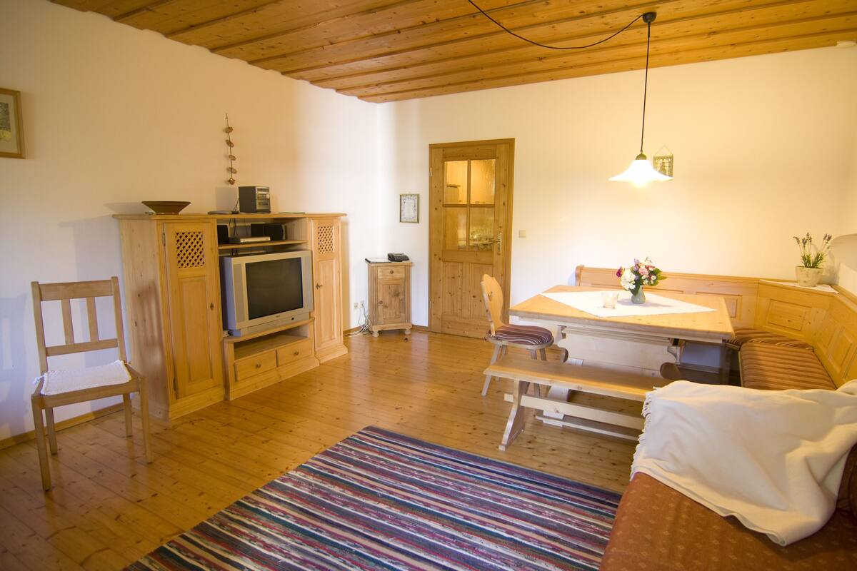 A cozy room with a wooden ceiling, a dining table with chairs, a sofa, a TV, and a wooden cabinet.