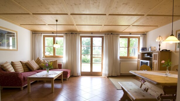 A cozy living room with a sofa, a coffee table, and a dining table with benches. There are large windows with curtains, a ceiling fan, and a lamp on the dining table.