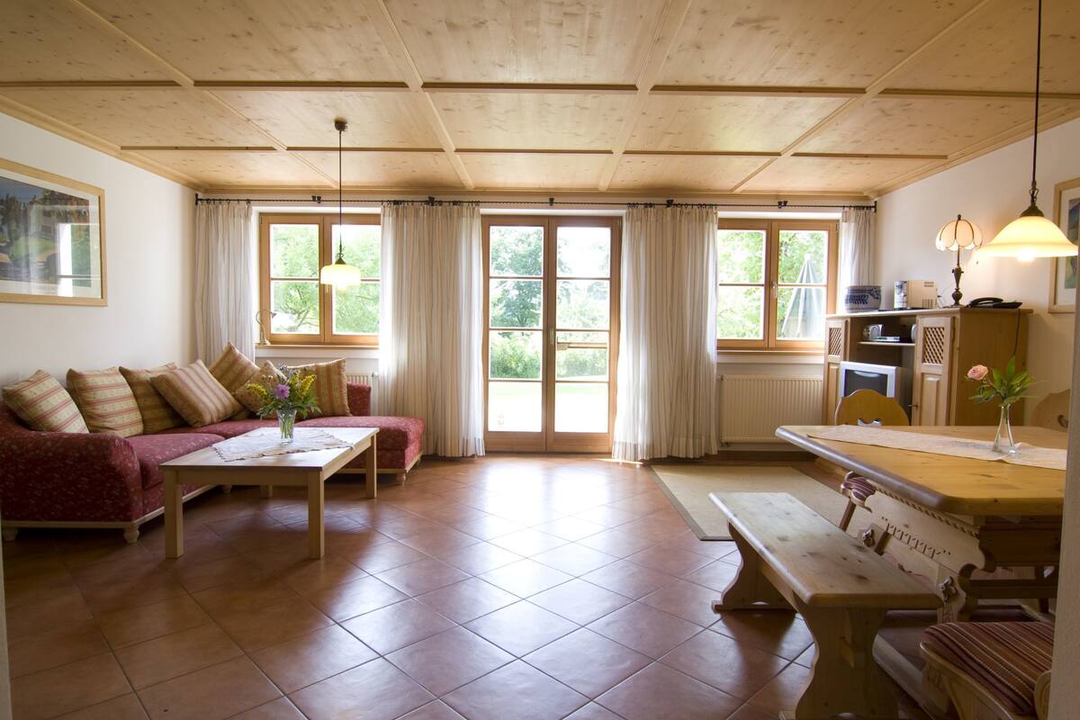 A cozy living room with a sofa, a coffee table, and a dining table with benches. There are large windows with curtains, a ceiling fan, and a lamp on the dining table.