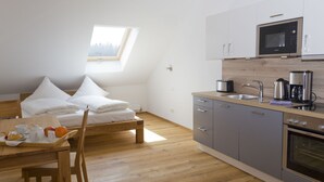 A compact kitchen with a microwave, coffee maker, and oven. There is a breakfast tray on a wooden table and a bed with pillows in the background.