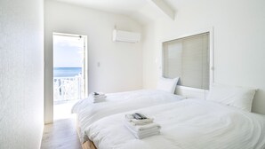 A bedroom with two beds, white linens, a view of the sea through a balcony door, and an air conditioning unit on the wall. - Villa Kanutan (Motobu)
