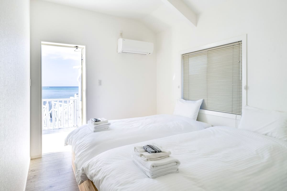 A bedroom with two beds, white linens, a view of the sea through a balcony door, and an air conditioning unit on the wall.