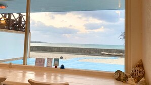 A beachside view from a window with a table and chairs, a boat, and a signboard. - Villa Kanutan (Motobu)