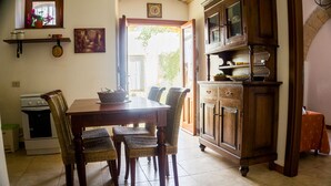 A dining area with a wooden table and wicker chairs, a kitchen with a stove and cabinets, and a view through an open door to the outside. - Agriturismo Castelluzzo (Custonaci)