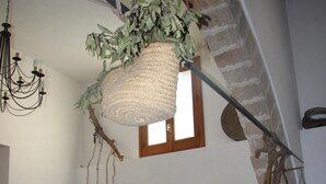 A spacious living room with a high ceiling, a white sofa, a hanging plant, a chandelier, and a staircase with exposed brick walls. - Agriturismo Castelluzzo (Custonaci)