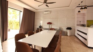 A modern dining area with a long table, brown chairs, a ceiling fan, and a kitchen area with white cabinets and a plant. - Seaview Winner Pool Villa (Ho Chi Minh City)